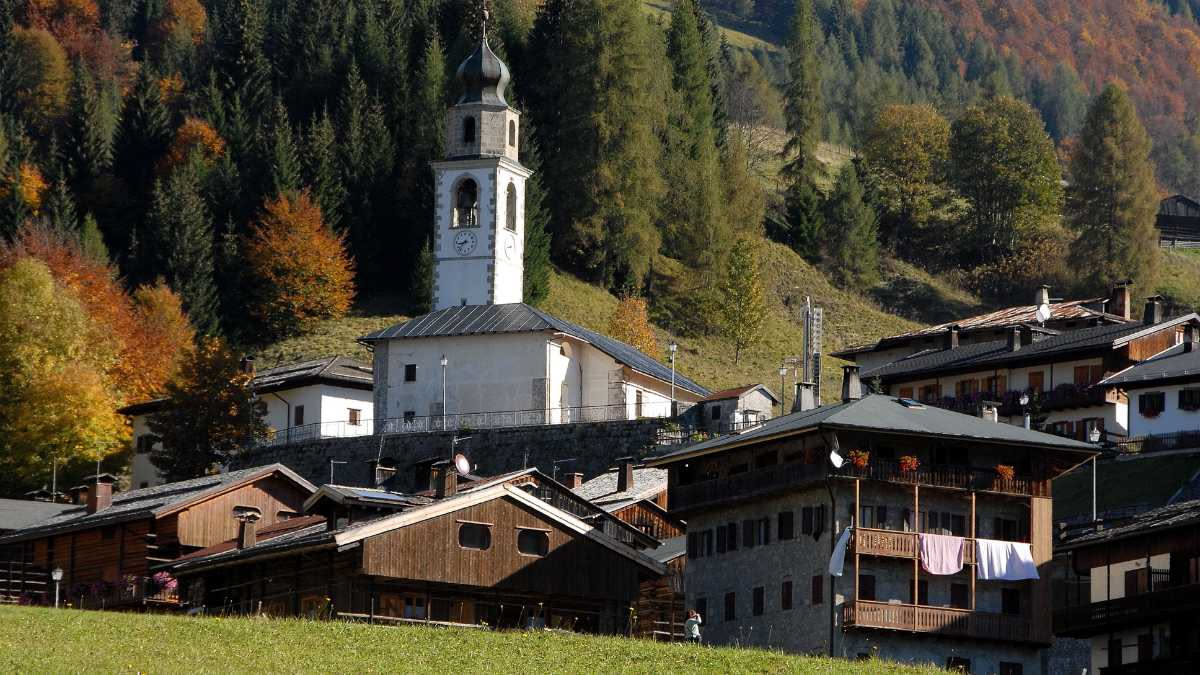Baita tradizionale sudtirolese in legno scuro con tetto sporgente rivestito di scandole, circondata da prati alpini verdi e picchi montuosi sullo sfondo
