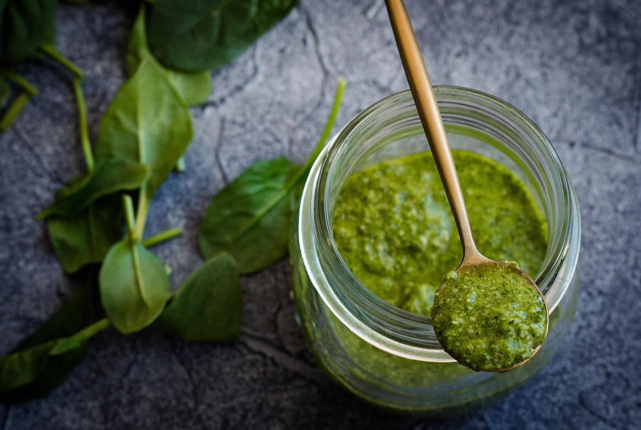 Tre vasetti di pesto genovese di marche diverse allineati su tavolo bianco, con etichette visibili e cucchiaio di legno accanto, basilico fresco sullo sfondo