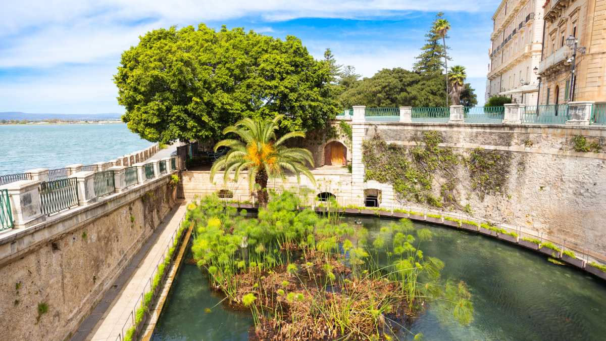 Giardino storico palermitano con aranci e limoneti nella Conca d'Oro, alberi centenari che creano ombra, fontana centrale circondata da vegetazione rigogliosa, vista della città sullo sfondo