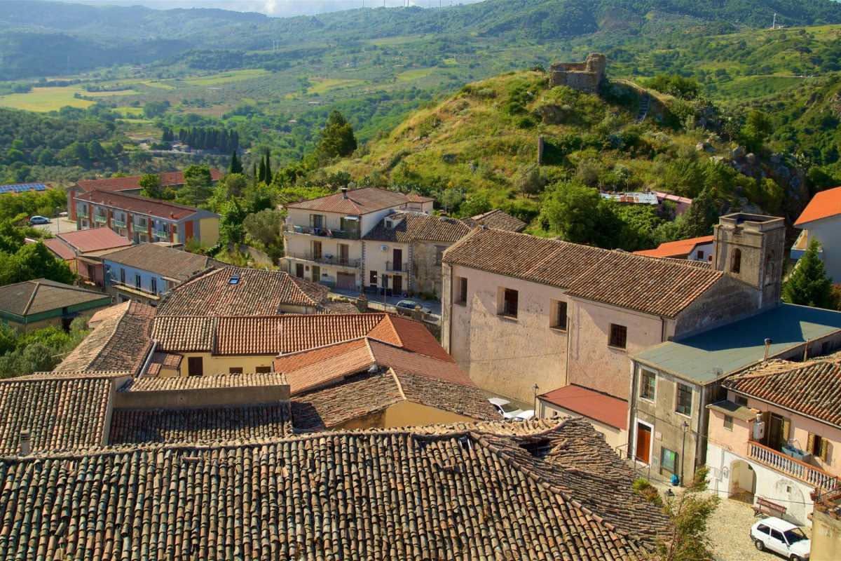 Piccolo agglomerato di case in pietra grigia e beige con muri spessi, finestre strette e porte di legno scuro in un paese collinare del Cilento; scale esterne di pietra e tetti di coppi rossi.