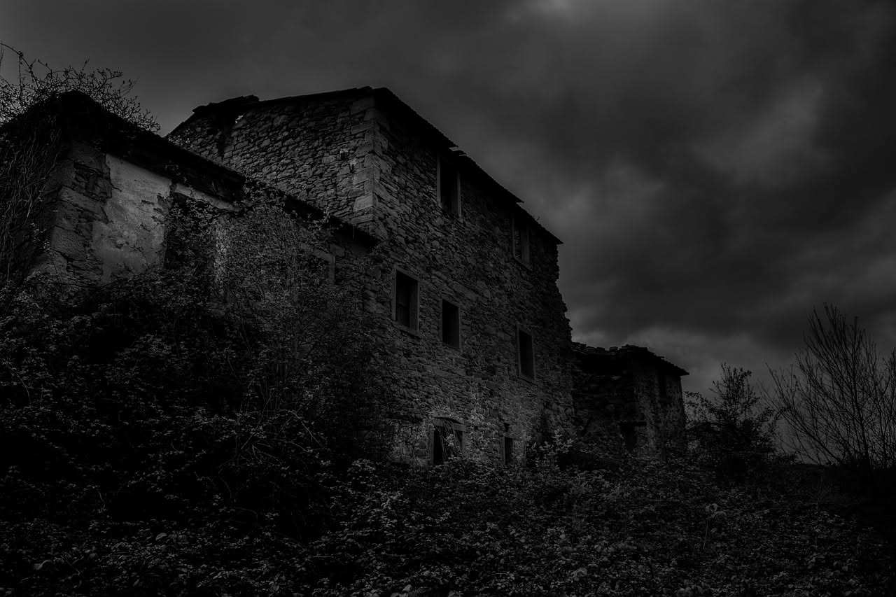 Parete di casa rurale calabrese con pietre scure e grigie a vista, malta grigia tra le fessure, finestra in legno invecchiato e arco in pietra naturale, vegetazione selvatica sulle pendici retrostanti