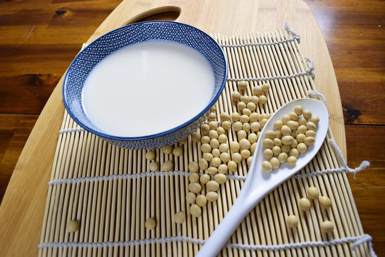 Due bicchieri di vetro trasparente, uno con latte bianco di soia e uno con latte beige di avena, con chicchi di soia e fiocchi di avena accanto su sfondo bianco