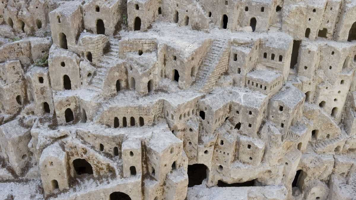 Facciata di casa in tufo grigio con crepe visibili, finestre rotte, vegetazione sulle mura, paesaggio collinare calabrese sullo sfondo