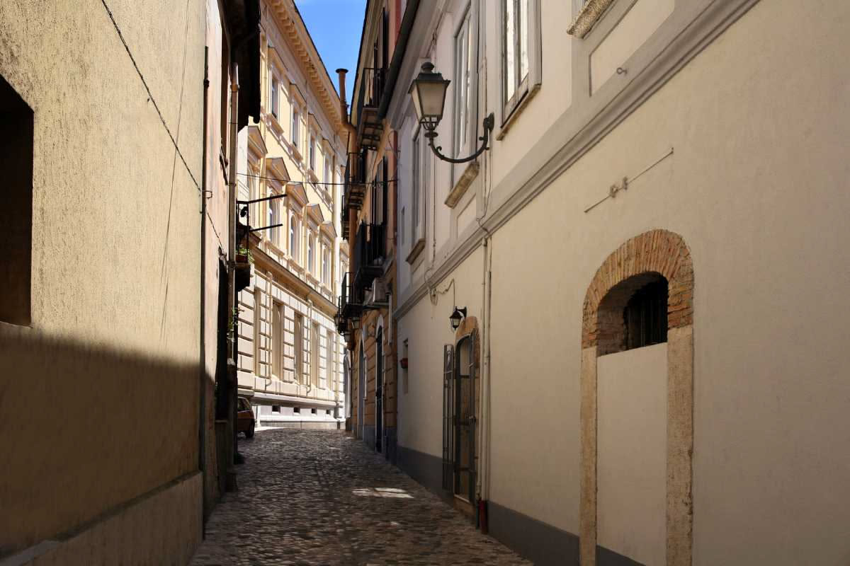 Lampione a gas in ferro battuto con braccio curvo e lanterna di vetro, installato su strada lastricata di una città italiana dell Ottocento, con edifici storici sullo sfondo