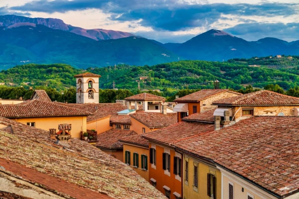 Scorcio di un borgo medievale italiano con tetti di tegole curve rosse che degradano verso vallata verde, montagne sullo sfondo, campanile in pietra che emerge tra le coperture
