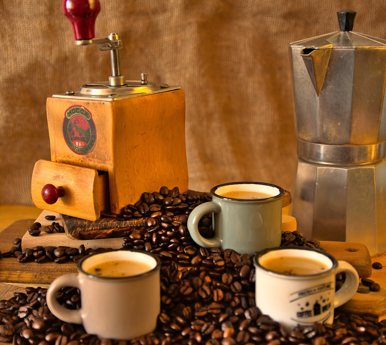 Chicchi di caffè marrone scuro e verde disposti su una tavola di legno, accanto a una tazza bianca e una misura di ottone per pesare