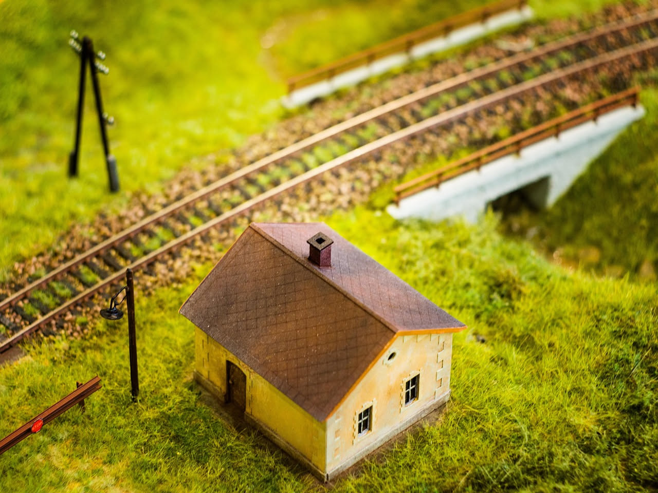Stazione ferroviaria di paese con casa bianca a due piani, tetto a doppia falda in coppi rossi, finestre simmetriche, binari paralleli sullo sfondo e alberi circostanti