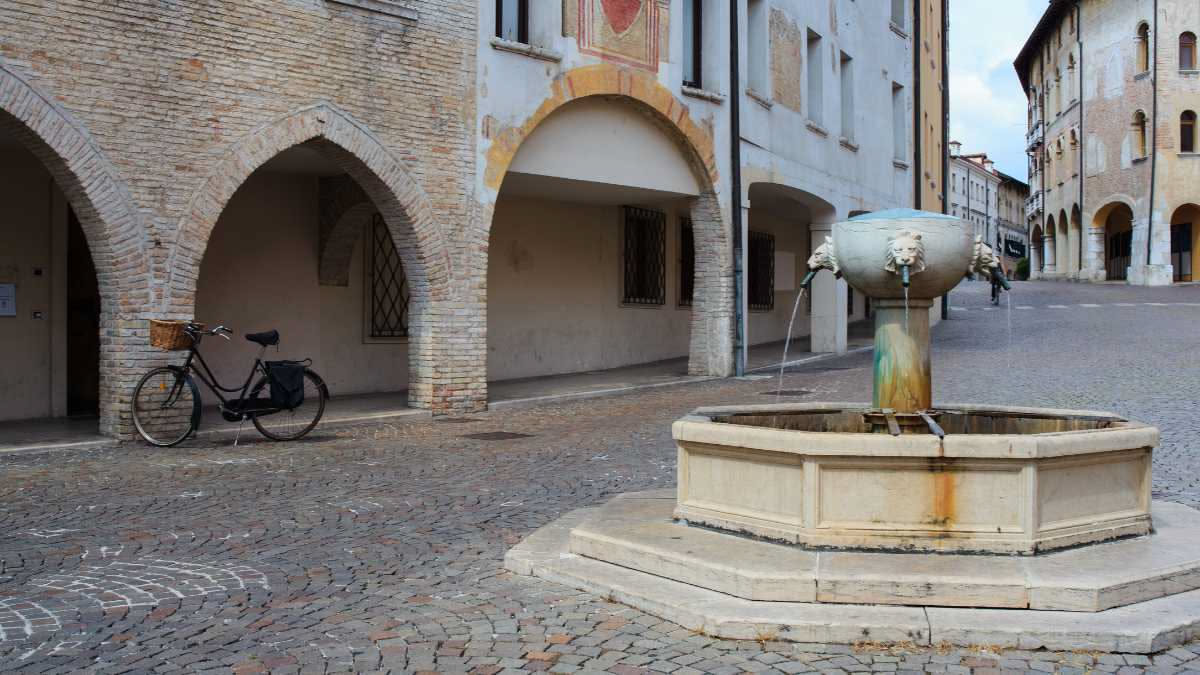 Fontana in pietra in piazza di un borgo medievale con tetti di coppi in primo piano e una cisterna sotterranea visibile attraverso una grata di ferro