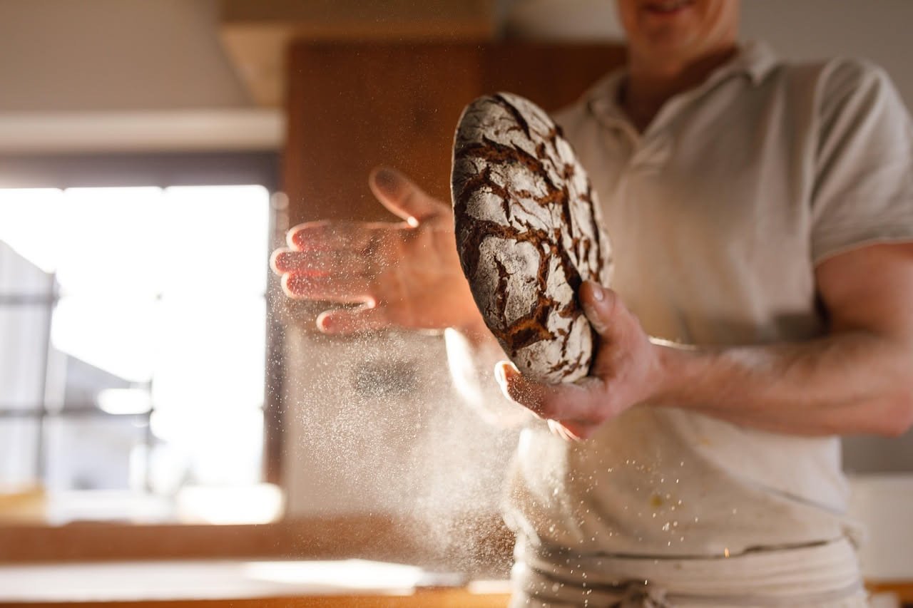 Pane rustico a lievito madre con crosta scura dorata, farina di grano locale dispersa intorno, mani di panettiere che lo taglia, sfondo di campo di grano verde