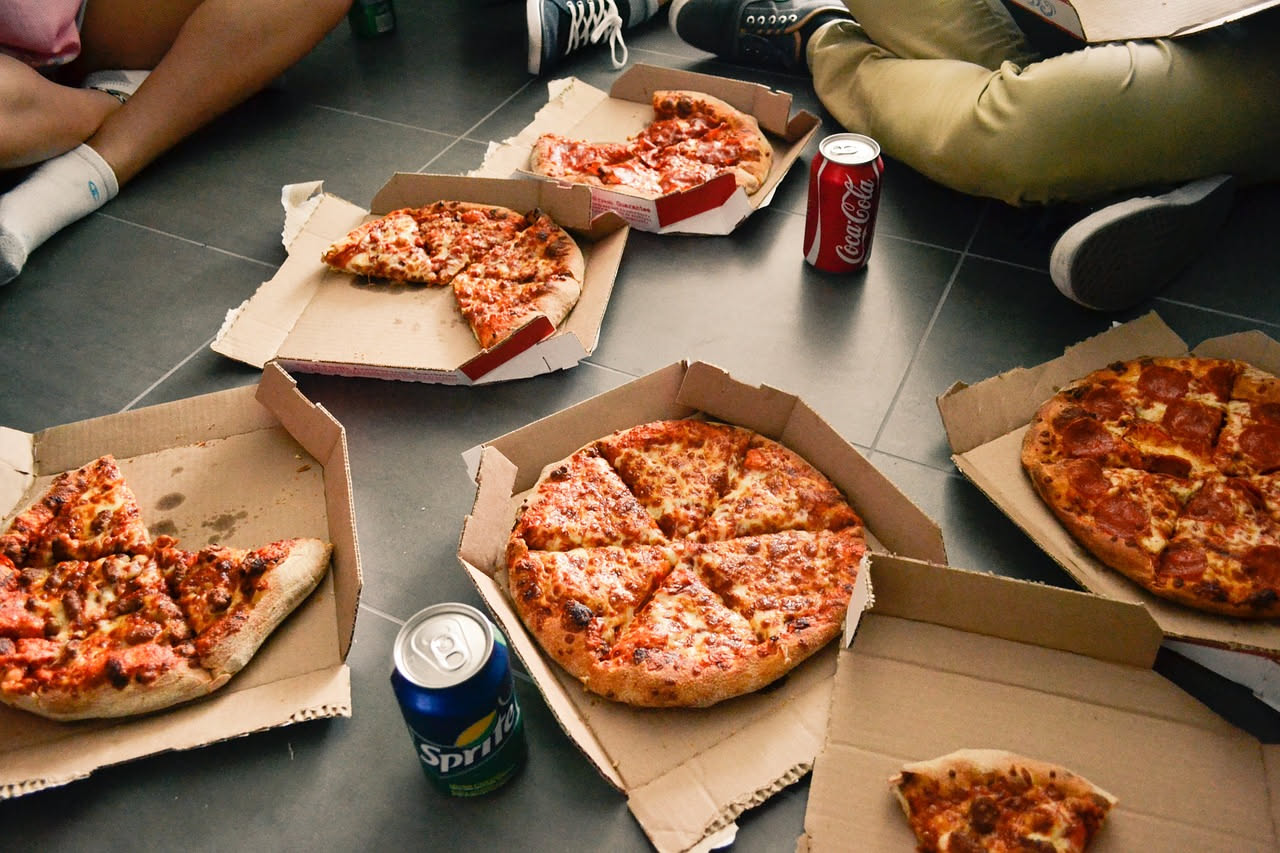 Tre cartoni di pizza puliti e appiattiti, disposti a ventaglio su una superficie bianca, accanto a forbici, spago naturale e pennarelli colorati per progetti di riciclo creativo