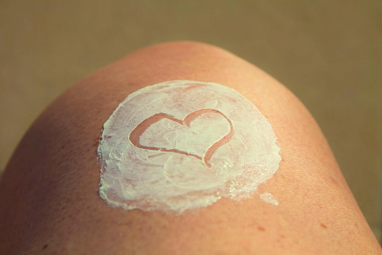 Mano che applica crema solare su braccio, con indumenti estivi di cotone bianco sullo sfondo e bottiglia di aceto trasparente accanto