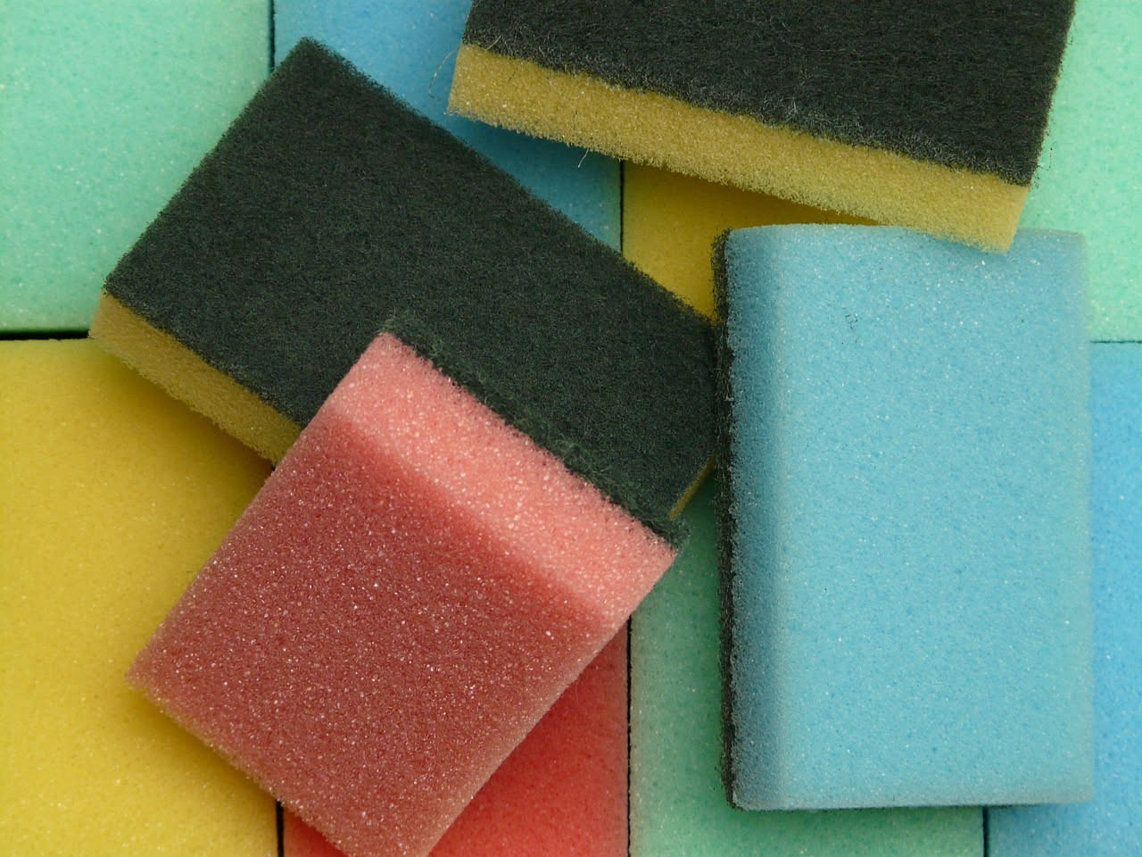 Spugne da cucina colorate su un piano di lavoro luminoso, accanto a una bottiglia di aceto bianco, un limone tagliato e un barattolo di bicarbonato di sodio