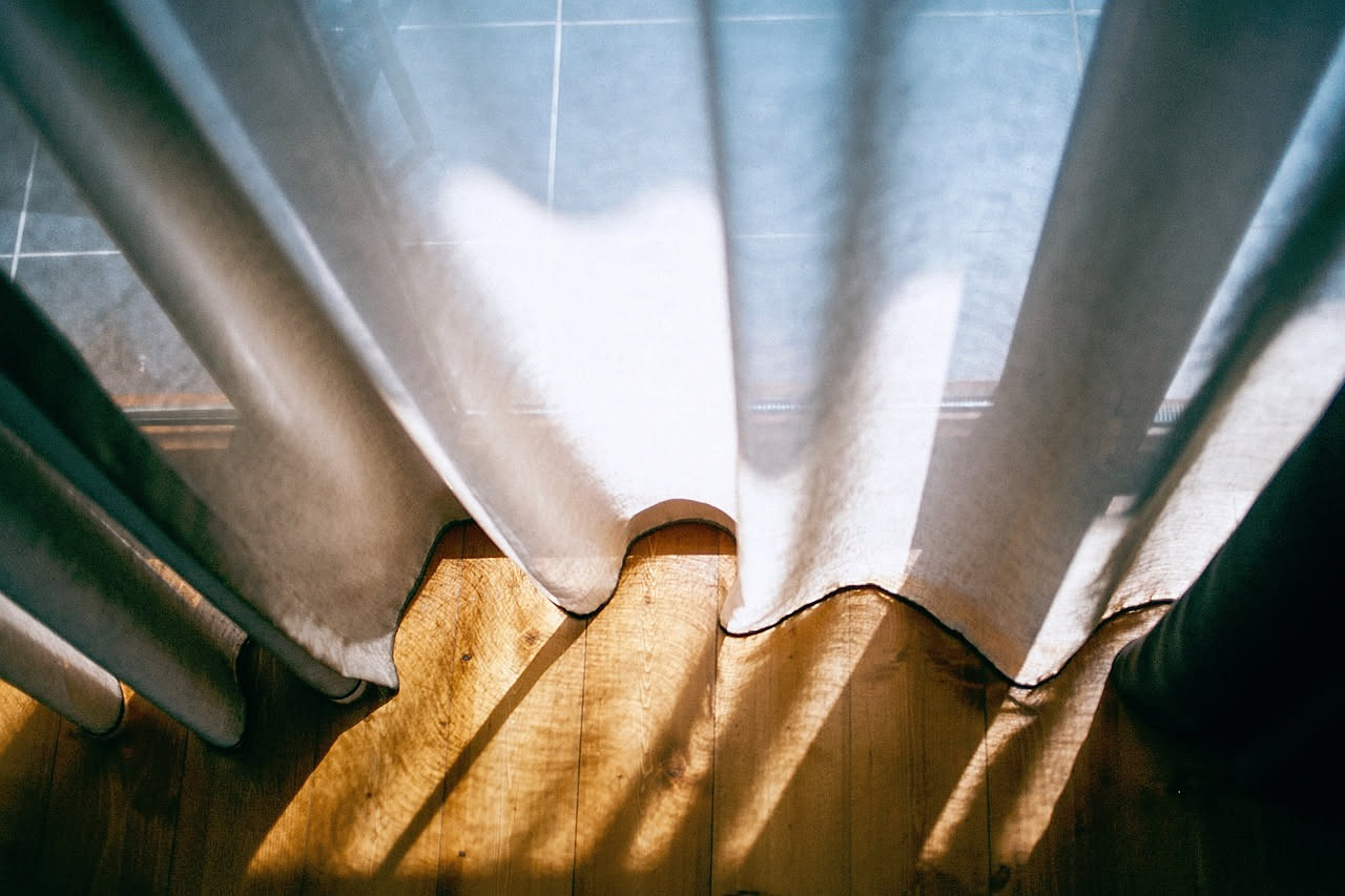 Tenda di mussola bianca che ondeggia leggera davanti a una finestra, la luce del sole la attraversa creando ombre dolci sulla parete