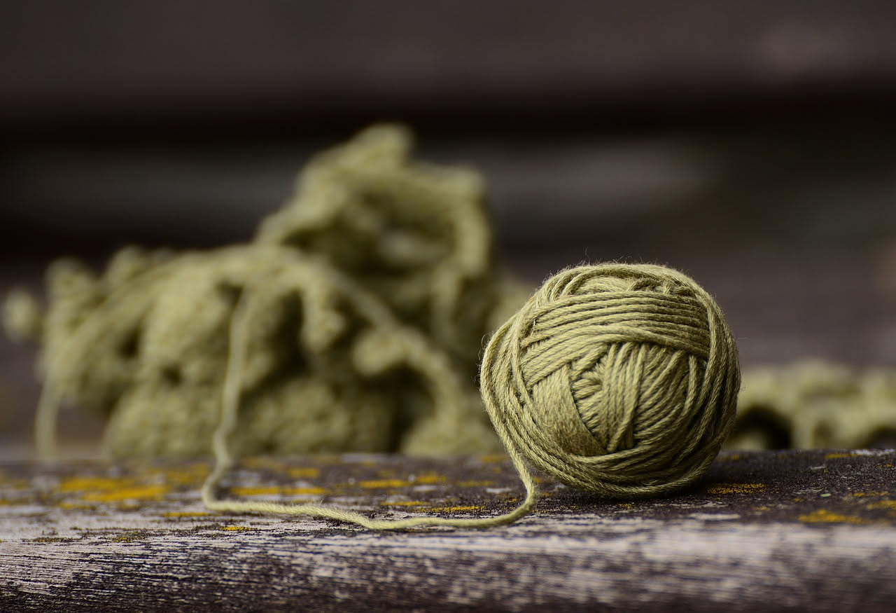 Ferri da maglia di diversi numeri e materiali, da legno a metallo, allineati accanto a gomitoli di filati di vari spessori, colori e fibre, su un tavolo luminoso.