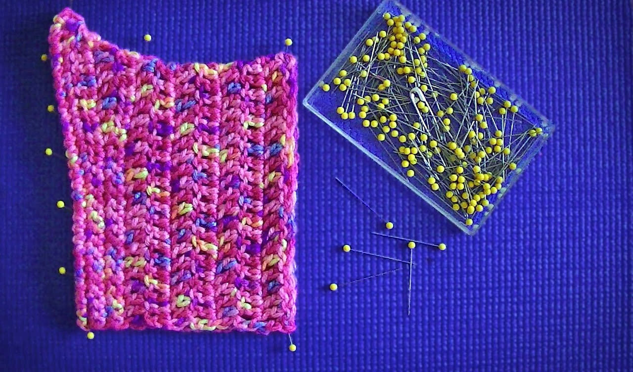 Uncinetto in metallo di varie misure accanto a gomitoli di filato colorato disposti su un piano di legno chiaro, con un lavoro a maglia in corso